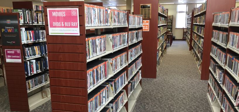 Shelves of DVDs appear in a public library. the shelves are bookended by mahogany, wooden panels. A sign on the end of a shelf reads "MOVIES, DVDS & BLU RAY"